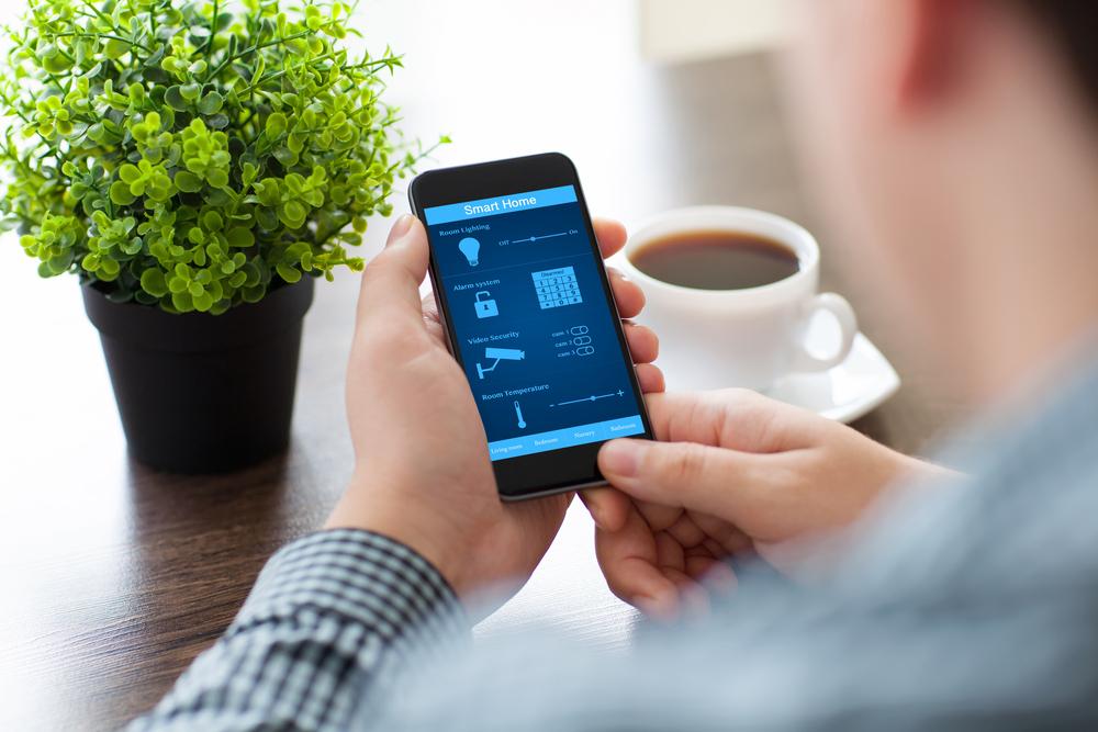 Person holding a smartphone displaying a Smart Home app, with a coffee cup and a potted plant on the table.